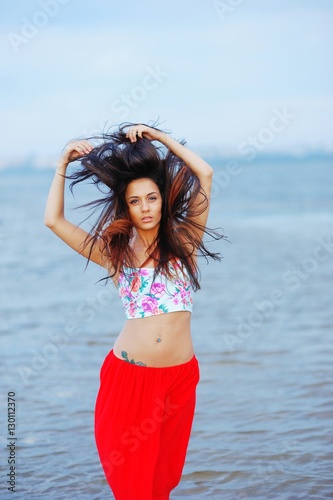 Portrait of beautiful slim woman, dressed in a t-shirt and red skirt at the hips, with piercings in navel. Girl on beach holding fluffy hair on blurred background of Gulf Finland