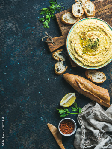 Homemade hummus dip in blue ceramic bowl with fresh baguette, lemon, herbs and spices over dark blue plywood background, top view, copy space