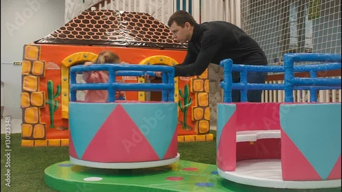 children round the carousel turns in the play area. Little girl climbs on it. Dad rolls his daughter