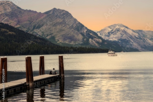 image of an evening sun setting behind the mountains reflecting on the lake with a man sitting on wooden pier and a boat sailing in the distance in the summer time.