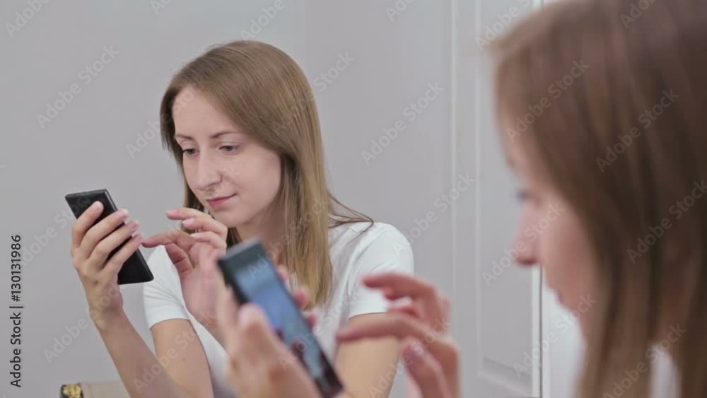 Pretty woman using smartphone sitting in front of mirror. White interior, makeup room Pretty woman using smartphone sitting in front of mirror. White interior, makeup room
