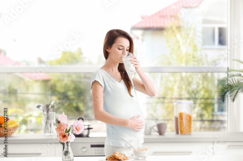 Young pregnant woman with glass of milk standing at kitchen