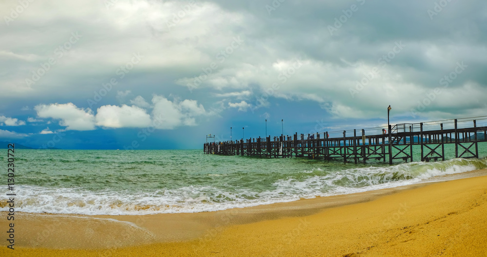 Old wooden pier stretching out to sea and foamy waves on the Maenam Beach, Koh Samui, Thailand. Rainy ominous grey storm clouds - dramatic sky