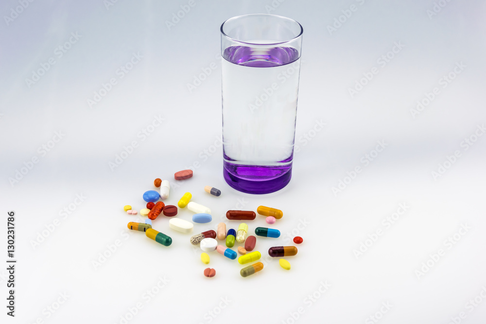 A lot og colored tablets and capsules near a glass of water with purple bottom isolated on white background.