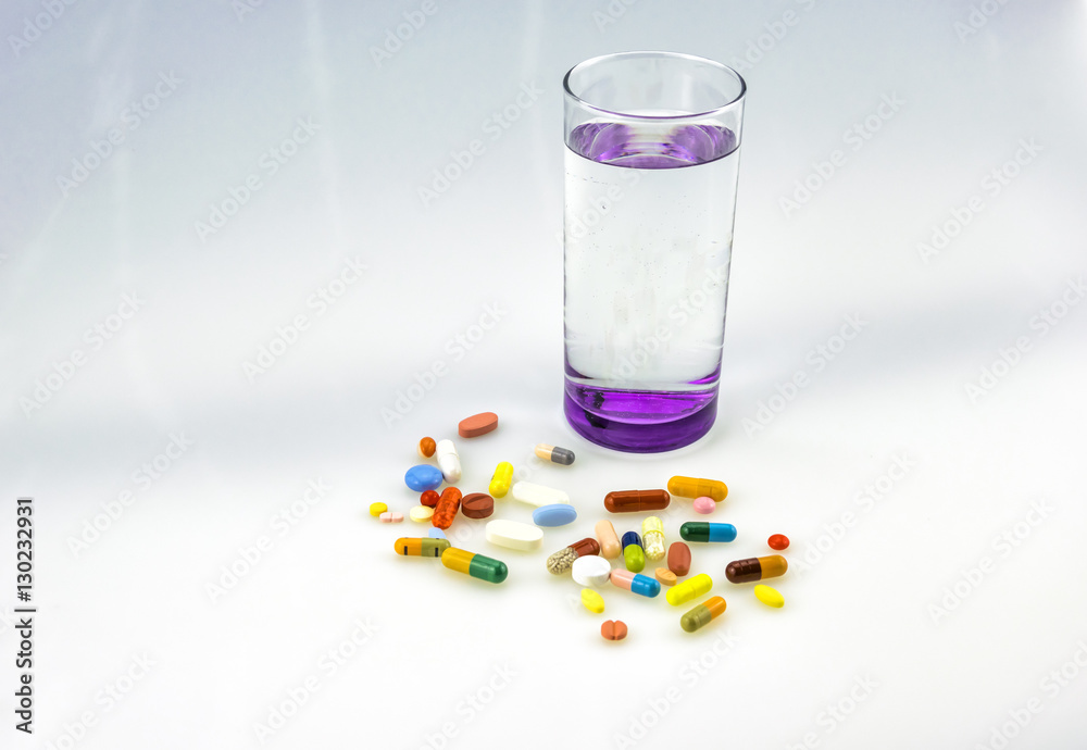A lot og colored tablets and capsules near a glass of water with purple bottom isolated on white background. A lot og colored tablets and capsules near a glass of water with purple bottom isolated on white background.