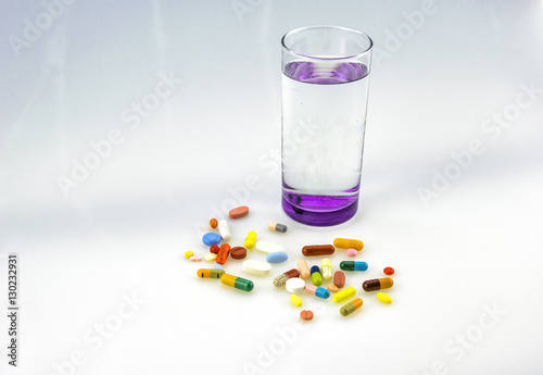 A lot og colored tablets and capsules near a glass of water with purple bottom isolated on white background.