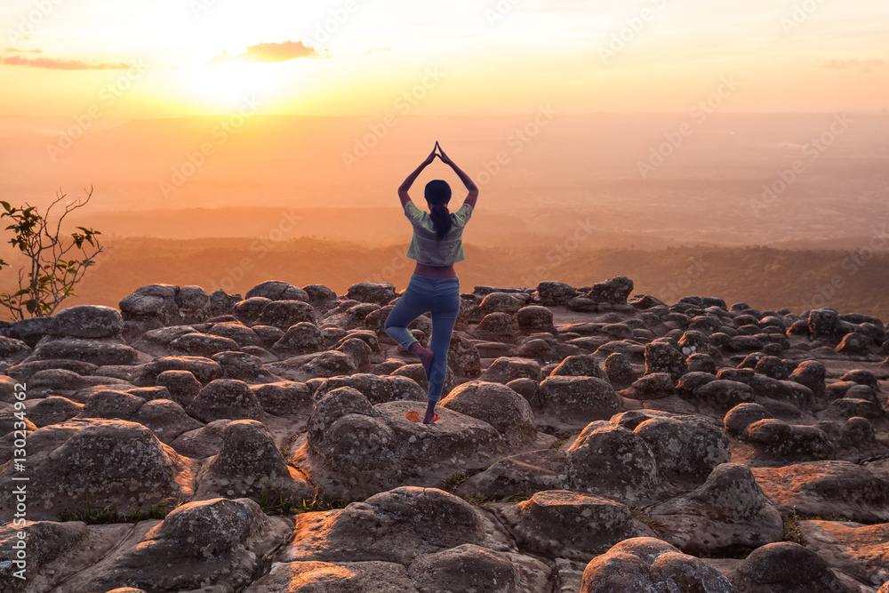 A woman practices yoga on silhouette of sunset and light flare at the mountain named Lan Hin Poom at Phu Hin Rong Kla National Park, Phitsanulok Province, Thailand