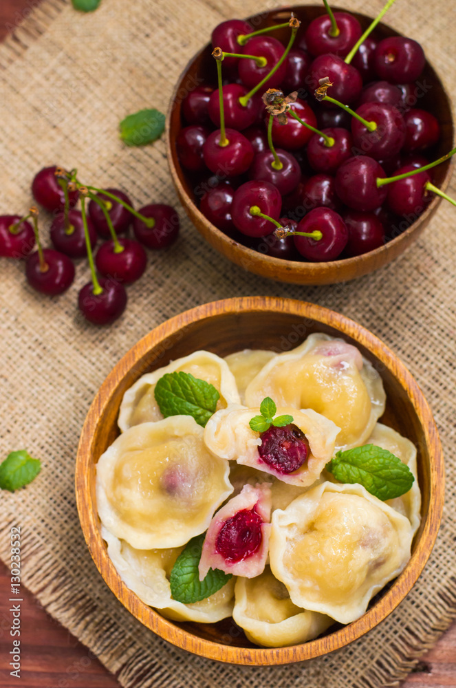 Dumplings with a cherry and mint leaves on background of flowers burlap. Wooden table. Top view. Close-up Dumplings with a cherry and mint leaves on background of flowers burlap. Wooden table. Top view. Close-up