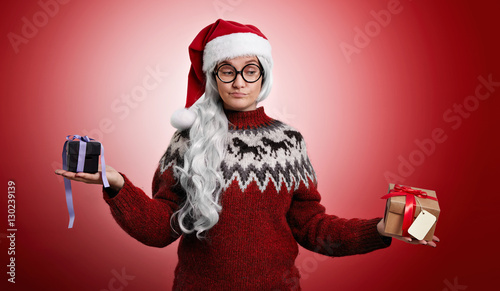 Undecisive woman in Christmas clothes and Santa hat weighing two presents in her hands isolated on red photo