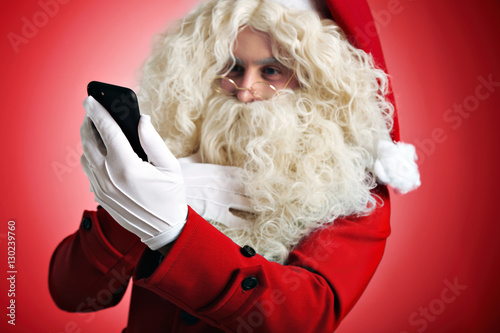 Exited Santa strokes his thick beard and looking in his mobile smart phone, isolated on red background photo