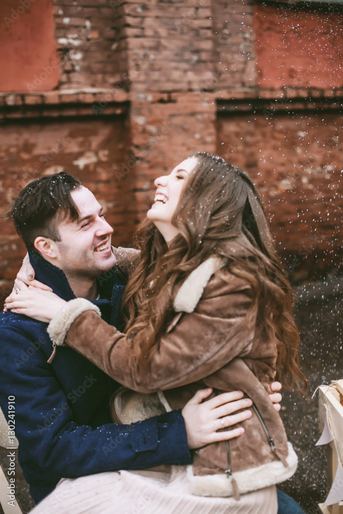 Young happy couple laughing and enjoying christmas street decorations with gifts in craft paper, cookies, cacao, handmade wooden table and red brick wall