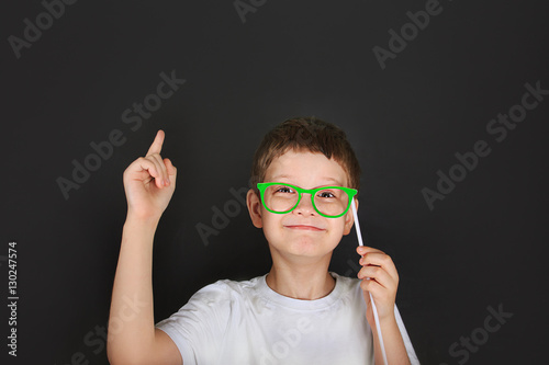 Smart boy with green glasses is thoughtful near chalkboard.