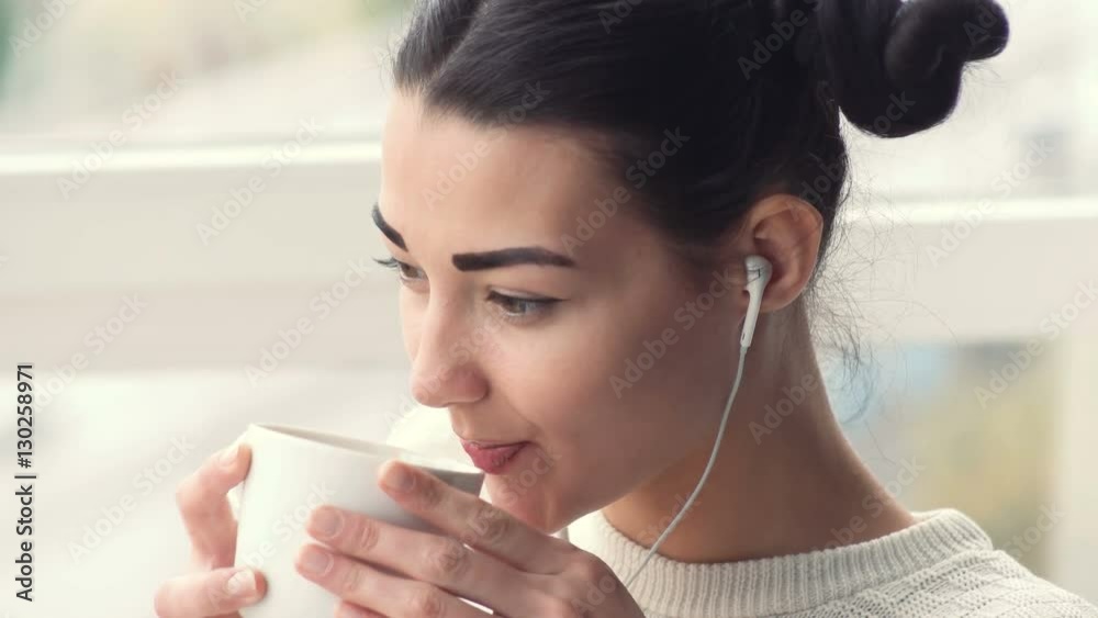 Charming young woman in earphones sitting near window listening musik and drink coffee Charming young woman in earphones sitting near window listening musik and drink coffee