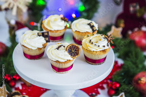 Пирожные, капкейки с сушеным лимоном и шоколадом на белой подставке на фоне новогодней зеленой гирлянды и новогодних огней.