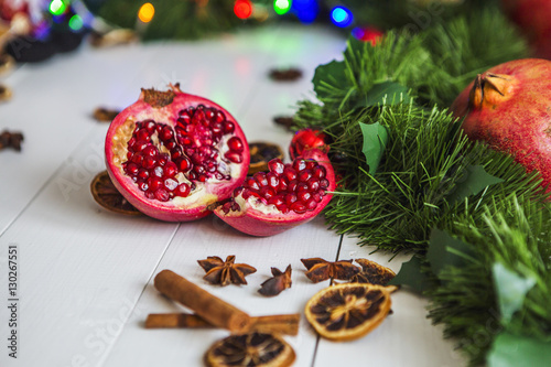 Разрезанный красный гранат, корица, сушеные лимоны лежат на белом деревянном столе на фоне зеленой новогодней гирлянды и новогодних огней.