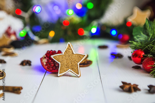Имбирный пряник в виде звезды, разрезанный красный гранат, корица, сушеные лимоны лежат на белом деревянном столе на фоне зеленой новогодней гирлянды и новогодних огней.