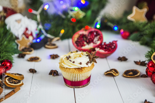 Пирожное с кремом, капкейк, мбирный пряник в виде звезды, разрезанный красный гранат, корица, сушеные лимоны лежат на белом деревянном столе на фоне зеленой новогодней гирлянды и новогодних огней.