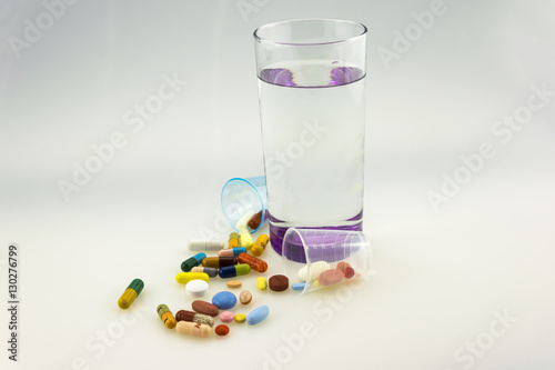 Many colored tablets and capsules near a glass of water with purple bottom isolated on white background.