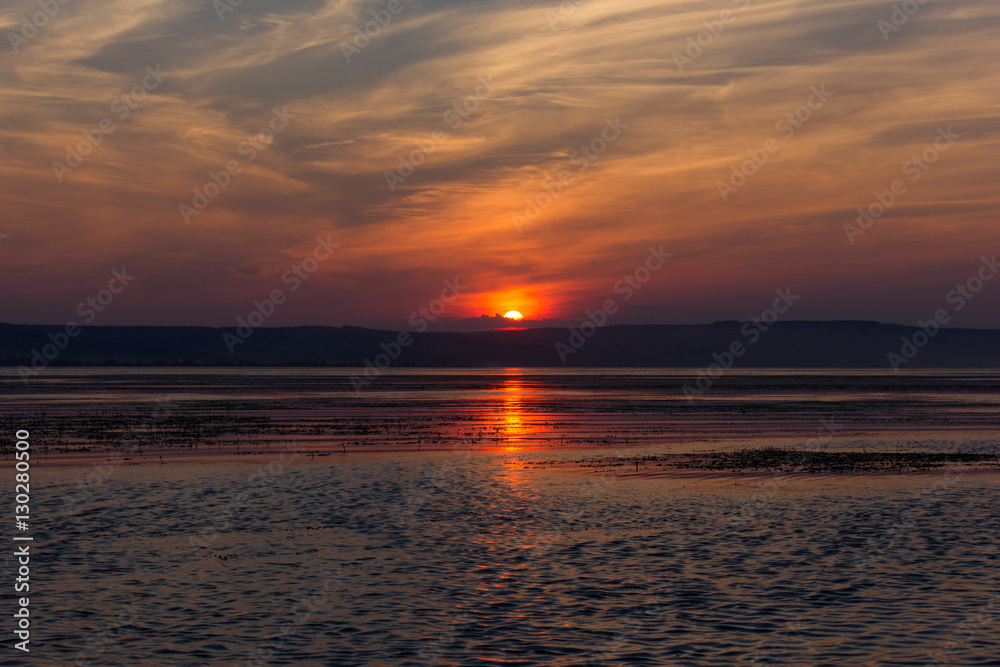 Red sunset over the water. Dramatic red sunset. Sun hiding behind the clouds at sunset, and reflected in the water.