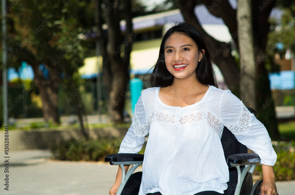 Naklejka premium Young brunette woman sitting in wheelchair smiling with positive attitude, outdoors environment, physical recovery concept
