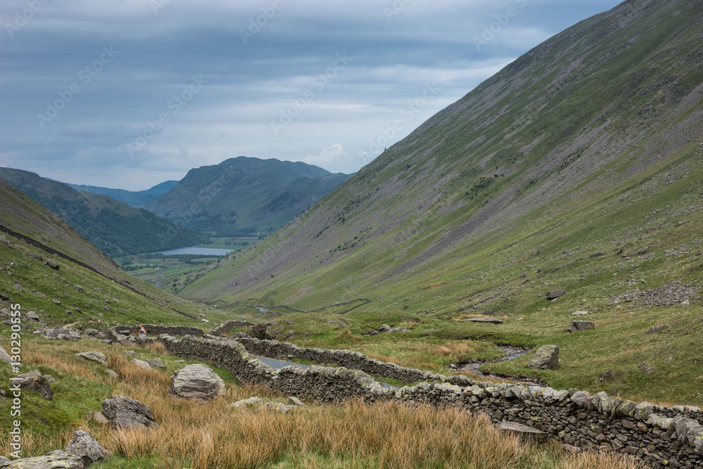 Fototapeta premium Lake District, England - May 30, 2012: Long rural road with stone walls on the side runs between two mountain slopes through the wide green landscape. Shot from hill. Lake at the horizon. Bluish sky.