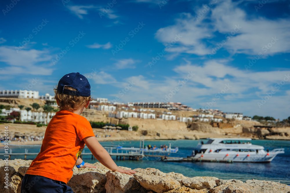 boy child is looking from the mountain to the beach with blue sea and nice boat yacht