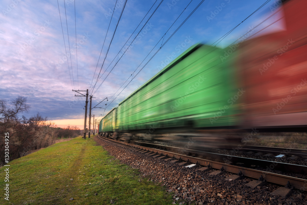 Fototapeta premium Railway station with cargo wagons and train in motion against blue sky with clouds at sunset. Colorful industrial landscape. Railroad. Railway platform. Heavy industry. Cargo shipping. Blurred wagons