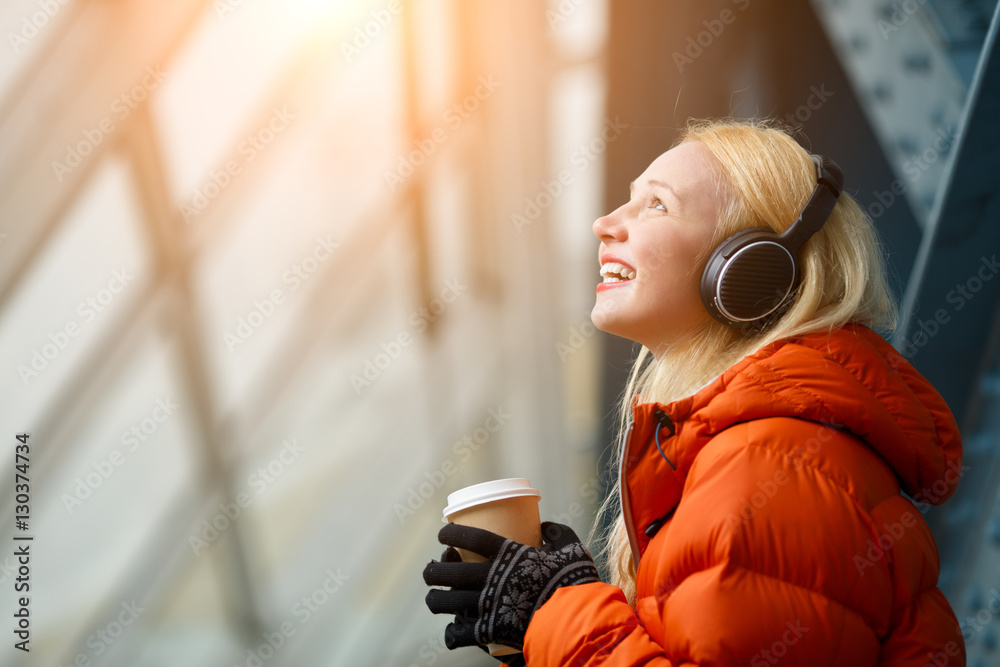 Happy young blonde woman in headphones holding coffee in hands