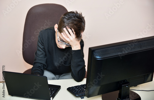 Tired young boy with glasses sitting and holding his head on the table between computer and laptop 