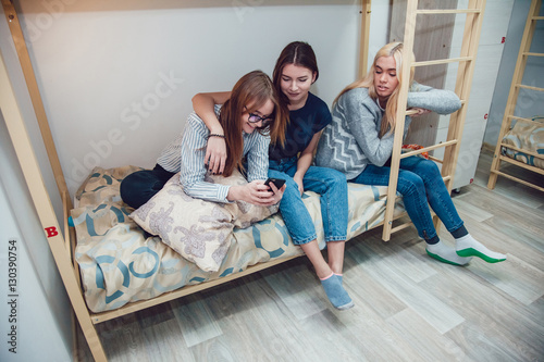 Canvas Print Beautiful girls sitting on a bed in hostel talk and using smartphone