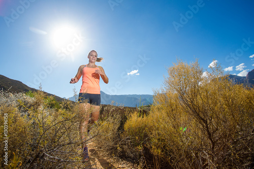 Wide angle view of a fit trail running female athlete running on a trail in the hills with dry shrubs on a bright sunny day