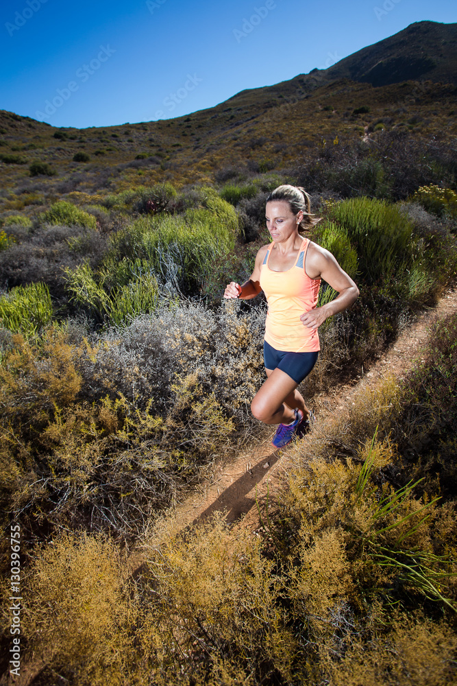 Wide angle view of a fit trail running female athlete running on a trail in the hills with dry shrubs on a bright sunny day