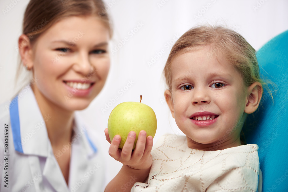 Cute girl holding green apple, sitting on chair in dental clinic. Concept of teeth cure and regular checkup. Cute girl holding green apple, sitting on chair in dental clinic. Concept of teeth cure and regular checkup.