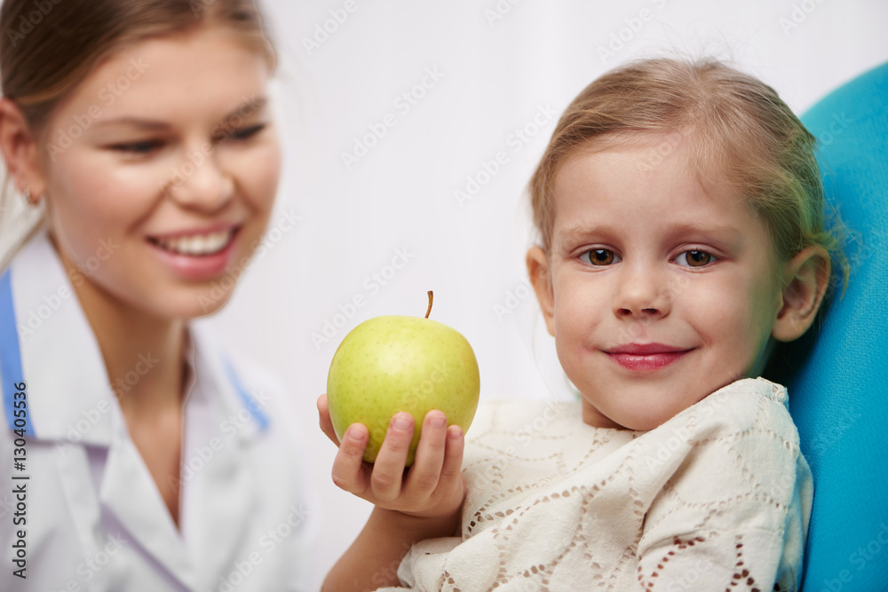 Milk teeth inspection. Smiling girl holding apple in dentist office.