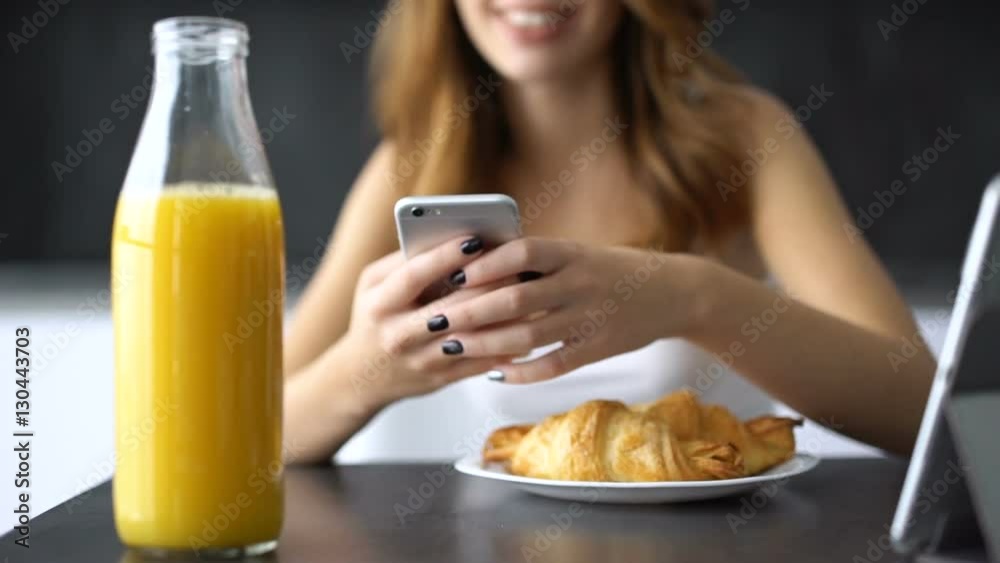 Happ young woman typing message on mobile phone while sitting on a kitchen and having breakfast Happ young woman typing message on mobile phone while sitting on a kitchen and having breakfast