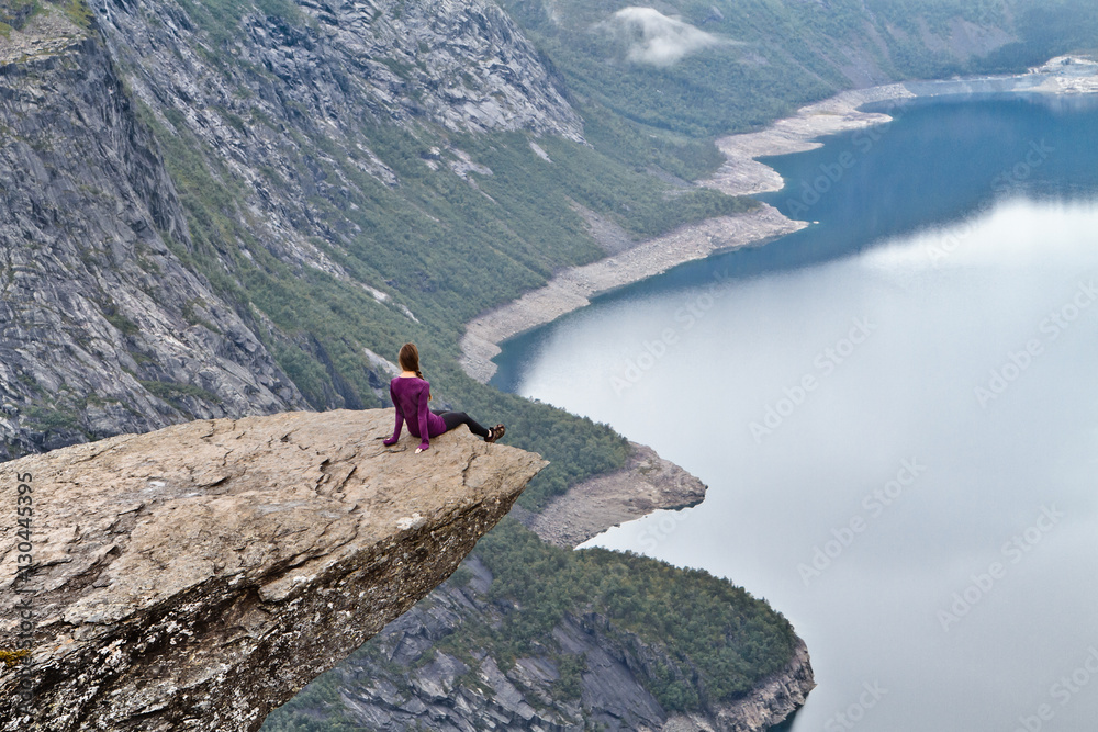 Girl sitting on Trolltunga rock (Troll's Tongue rock) and looking at Norwegian mountain landscape