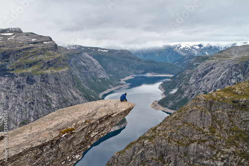 Man sitting on Trolltunga rock (Troll's Tongue rock) and looking at Norwegian mountain landscape