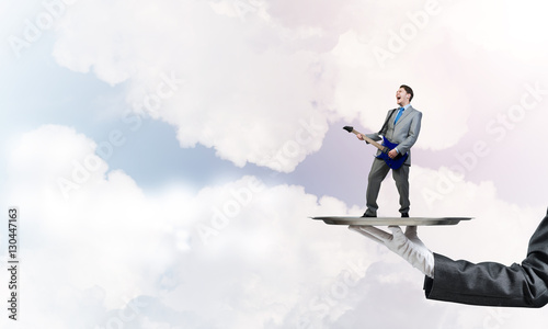 Businessman on metal tray playing electric guitar against blue sky background