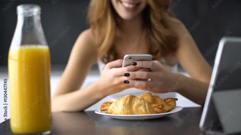 Cheerful smiling young woman texting message on mobile phone while sitting on a kitchen and having breakfast Cheerful smiling young woman texting message on mobile phone while sitting on a kitchen and having breakfast