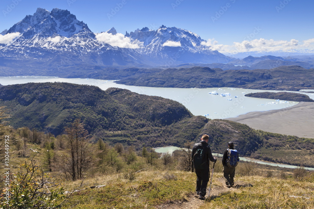 Hikers descend a grassy slope with lake, mountain and iceberg view; near Ferrier Vista Point, Torres del Paine, Patagonia, Chile Hikers descend a grassy slope with lake, mountain and iceberg view; near Ferrier Vista Point, Torres del Paine, Patagonia, Chile