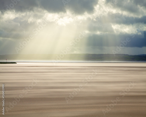 Crepuscular rays through a stormy sky while shifting sands create a cloud underfoot as wind whistles across the beach, West Kirkby, Wirral