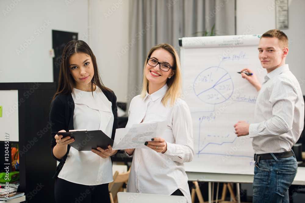 Two beautiful office workers having conversation while man colleague drawing business strategy on flip chart. Two beautiful office workers having conversation while man colleague drawing business strategy on flip chart.