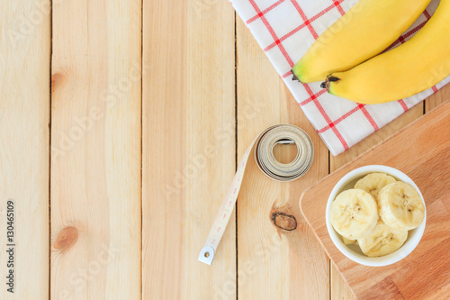 Two bananas and banana slices in white bowl with measuring tape on wooden table background, Top view with copy space and text.