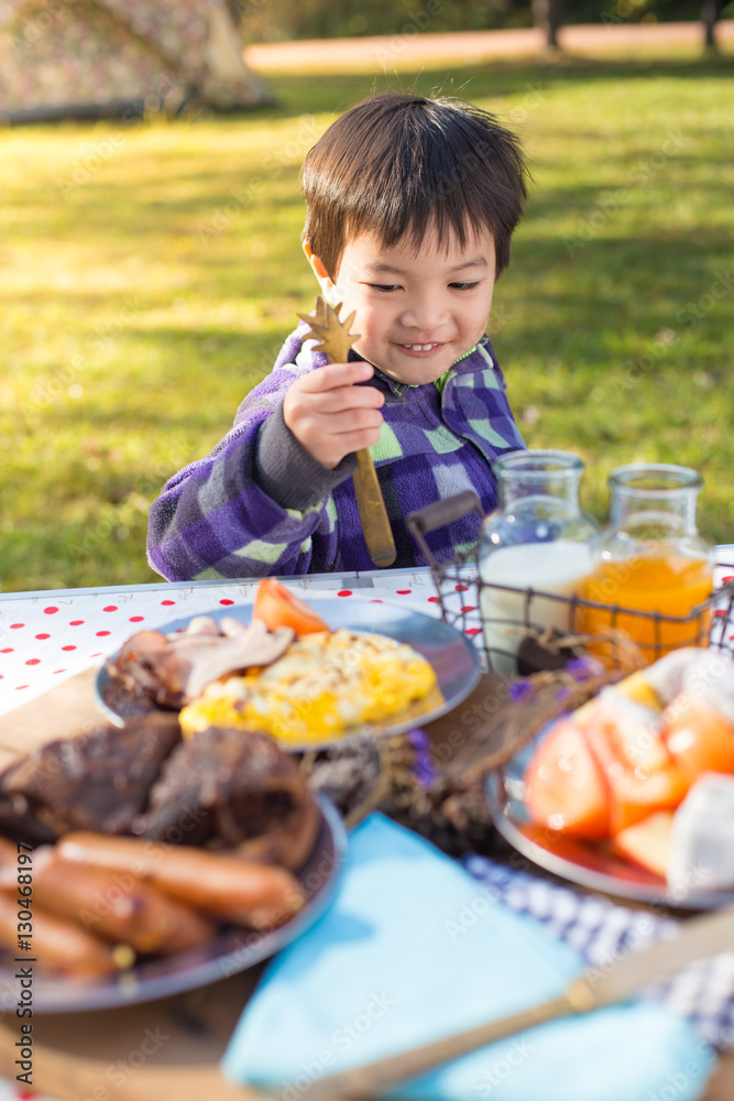 An Asian cute young boy eating breakfast outdoor at camping site in nature sunlight with a tent at the background. An Asian cute young boy eating breakfast outdoor at camping site in nature sunlight with a tent at the background.