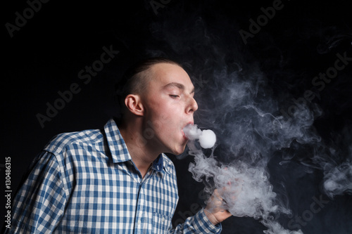 A young man with a stylish haircut in a blue plaid shirt smokes a cigarette elektronnuju on a black background. Exhaust fumes from cigarettes makes the rings figures