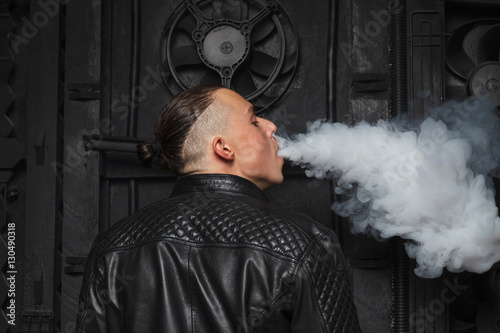 Fashionable and stylish man standing on black background with details and smoke E-cigarette in a leather black jacket, gray smoke releasing