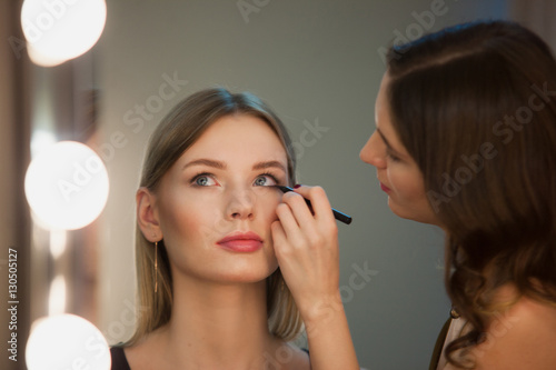 Makeup artist applies makeup to attractive blonde girl at the mirror with lamps in the beauty studio.