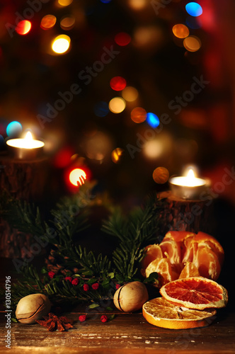 dark picture of the situation of winter holiday. Dried orange slices  berries  nuts and burning candles on a brown wooden background  blurred focus outcomes decorated Christmas tree  