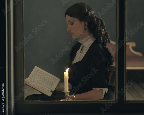 Obraz na plátně Retro victorian girl reading book by candlelight behind window.