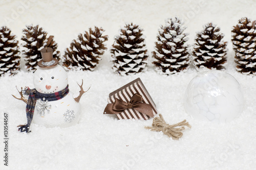 Gift box and snowman are in the snow. Christmas light background has cones. 